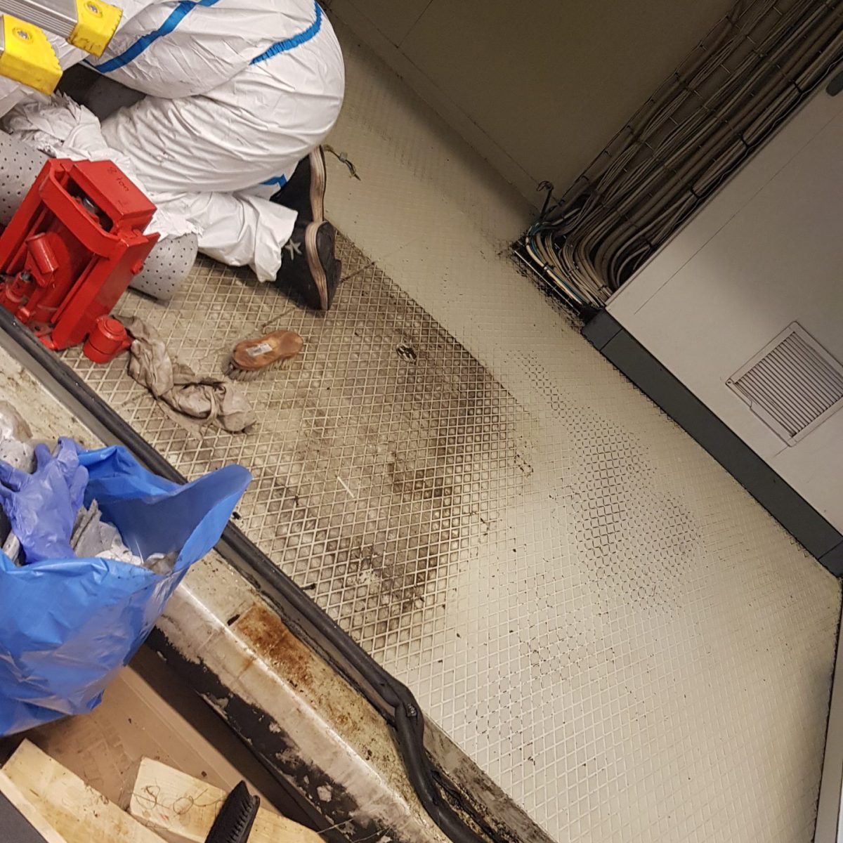 En person i skyddsdräkt och med arbetshandskar är böjd över och arbetar på golvet i ett rum med teknisk utrustning. Rummet har många kablar längs väggen och golvet är täckt av små, vita plattor. Det finns skräp och arbetsverktyg, som en hammare och träblock, utspridda på golvet. Situationen antyder en renovering eller reparation av golvet eller underliggande installationer.