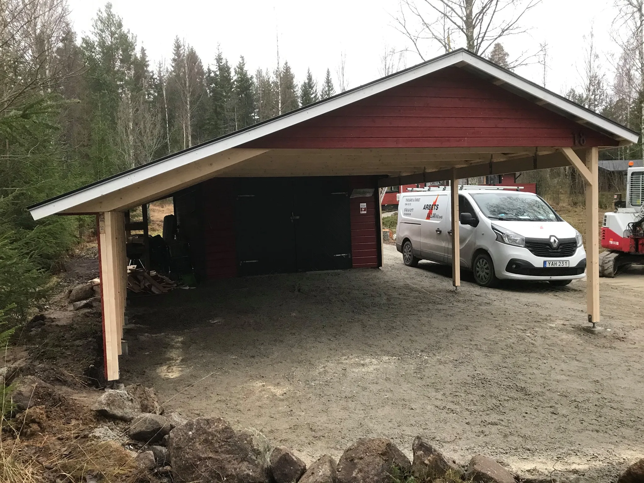 Ett rött träfärgat carport med ett vitt tak som skyddar en vit arbetsbil. Bakom carporten syns en barrskog och området runt är gruslagt. På bilen syns texten 'Arbetskällan'. Till vänster i bilden syns en öppen svart dörr som leder till ett förrådsutrymme under carporten, där diverse föremål lagras