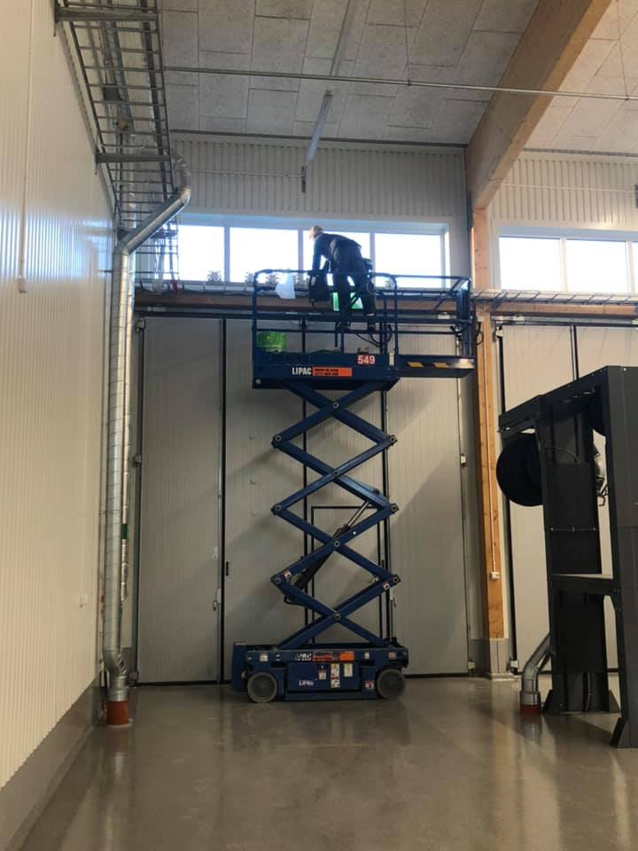 En arbetare står på en upphöjd, blå saxlift inuti en industrihall för att utföra arbete på en hög nivå. Hallen har metallväggar och ett högt tak med synlig ventilation och belysning, vilket antyder arbete på en industri- eller lagerbyggnad.