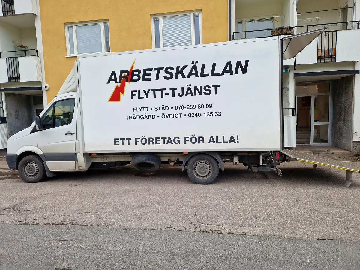 En vit flyttbil parkerad framför en gul flerfamiljsbyggnad. På sidan av lastutrymmet finns företagsnamnet 'ARBETSKÄLLAN FLYTT-TJÄNST' i stora bokstäver, med en logotyp som avbildar en röd blixt. Det finns telefonnummer för kontakt: 'FLYTT - STÄD - 070-289 89 09' och 'TRÄDGÅRD - ÖVRIGT - 0240-135 33'. Nedanför dessa står det 'ETT FÖRETAG FÖR ALLA!'. Bakdörren till lastutrymmet är öppen, och en utfällbar ramp är synlig.