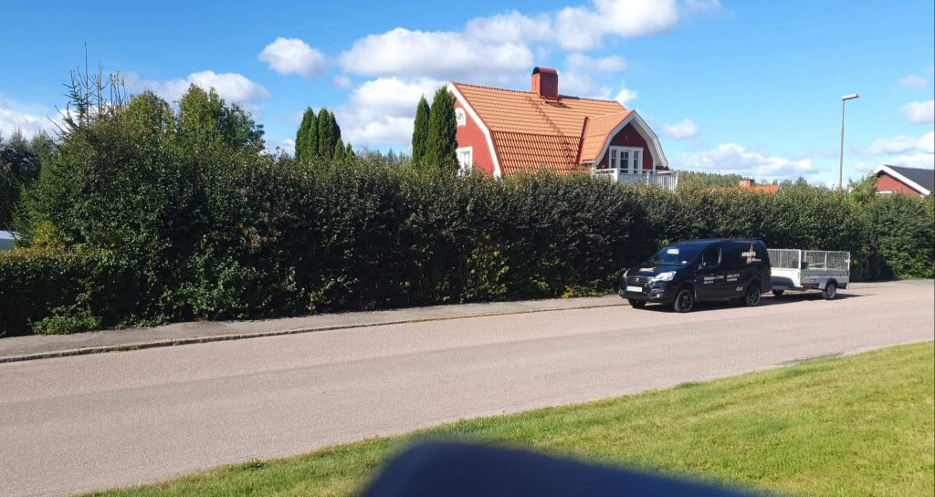 Bilden visar en idyllisk svensk villa med röda tegelväggar och ett mörkt tak, skyddad av en tät häck. Framför huset syns en arbetsbil med släp och en klarblå himmel ovanför, vilket antyder en solig och klar dag.