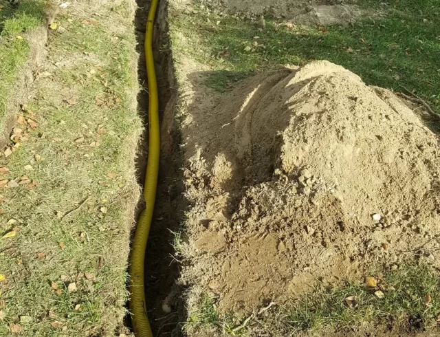 Bilden visar en grävd ränna i marken där gula flexibla rör eller slangar har lagts ner. Det ser ut som att det kan vara en installation av någon typ av underjordiskt system, möjligtvis för dränering eller bevattning. Jorden runt rännan har grävts upp och ligger i högar längs med grävningen. I bakgrunden syns grönska och några löv som börjar ändra färg, vilket kan tyda på att det är höst. Det finns inga synliga verktyg eller personer i bilden, så det är oklart om arbetet är avslutat eller om det kommer att fortsätta.